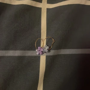 Två par färgglada örhängen i guld - Säljer två par örhängen med guldfärgade hoops. Ena paret har blommor i lila och ljusblått, det andra paret har pärlor och stavar i blått, lila och rosa. Perfekta för dig som gillar unika och lekfulla accessoarer!