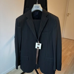 Svart kostym – Rice, Made in Italy (stl 46) - Säljer en helt ny och oanvänd kostym från italienska märket Rice, tillverkad i Italien. Klassisk svart design med diskreta vertikala ränder – perfekt för både arbete, fest och mer formella tillfällen.  Detaljer:  Märke: Rice  Storlek: 46  Färg: Svart med subtila ränder  Tillverkad i: Italien  Skick: Ny med taggar kvar  Välskräddad med dubbla framfickor, bröstficka och innerfickor. Väldigt bekväm passform med fin