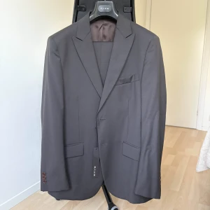 Svart/mörkbrun kostym – Rice, Made in Italy (stl 50) - Helt ny kostym från italienska Rice. Färgen är en elegant mörk nyans – svart med dragning åt mörkbrun, vilket gör den stilren men samtidigt lite annorlunda än en klassisk svart. Tvåknäppt modell med matchande byxor och brunt sidenfoder med vinröda detaljer.  Storlek: 50  Färg: Svart/mörkbrun (col. 9)  Märke: Rice – Made in Italy  Nypris: 4495 kr  Skick: Ny med tags