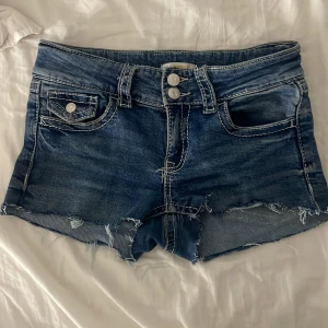 Blå jeansshorts från Gina Tricot - Snygga blå jeansshorts från Gina Tricot med råa, fransiga kanter och dubbla knappar framtill. Klassisk femficksmodell med kontrastsömmar och slitna detaljer för en cool look. Perfekta för varma dagar och enkel att matcha med allt.