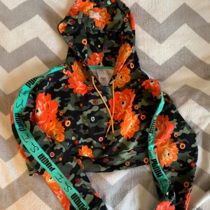 Puma x Sue Tsai camo hoodie med blommor - Säljer en unik hoodie från Puma x Sue Tsai i storlek M. Tröjan har camouflagemönster i grönt och svart, stora orange blommor och färgglada detaljer. Ljusgula snören och turkosa band med Puma-tryck längs ärmarna. Perfekt för dig som gillar statement-plagg.