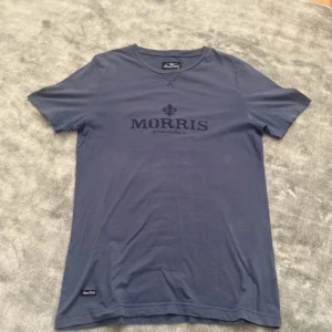Blå t-shirt från Morris Stockholm - Snygg blå t-shirt från Morris Stockholm i storlek medium. Klassisk rund hals och korta ärmar, med tryckt logga framtill. Skön passform och mjukt material som känns chill att bära. Perfekt för dig som gillar stilrena plagg med diskret branding.