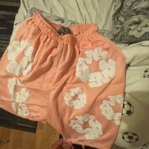 Rosa  shorts från Denim Tears - Säljer ett par peachfärgade shorts från Denim Tears med vita blommiga tryck över hela plagget. De har elastisk midja för skön passform och är perfekta för varma dagar. Riktigt snygga och unika shorts som sticker ut i mängden.