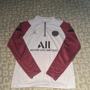 PSG långärmad träningströja Jordan - Snygg långärmad PSG träningströja i vitt med vinröda ärmar och svarta detaljer. Tröjan har guldigt Jordan-logga och PSG-märke på bröstet. Materialet är lätt och andas, perfekt för fotbollsträning eller chill. Halv dragkedja framtill för extra stil.