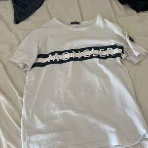 Vit Moncler t-shirt med logga - Vit t-shirt från Moncler med svart och vit rand över bröstet och stor 3D-logga. Klassisk rund halsringning och korta ärmar. Moncler-märke på ena ärmen. Perfekt för dig som gillar stilrena och exklusiva plagg.