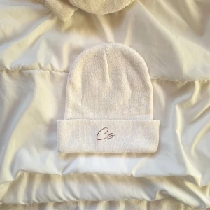 Vit stickad mössa med Cs brodyr - Snygg vit stickad mössa med uppvikt kant och stilren Cs-brodyr framtill. Enkel och clean design som passar till allt, perfekt för dig som gillar minimalistisk stil. Mjuk och skön kvalitet som håller dig varm under kyliga dagar.