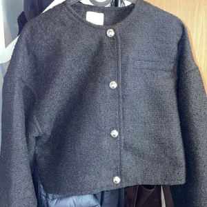 Svart cardigan jacka från H&M - Svart cardigan jacka från H&M med grovstickad känsla och snygga silvriga knappar framtill. Jackan är croppad och har rund halsringning samt en diskret bröstficka. Perfekt för dig som gillar stilren och enkel design.