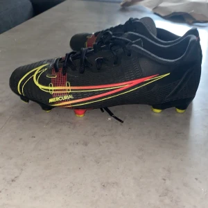 Nike Mercurial svarta fotbollsskor - Säljer ett par svarta Nike Mercurial fotbollsskor med snygga gula och röda detaljer. Skorna har snörning, låg profil och mönstrad ovandel i syntetmaterial. Perfekta för dig som vill sticka ut på planen med stilrena och sportiga skor.