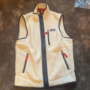 Vit fleeceväst från Patagonia XS helt ny  - Retro pile fleeceväst från Patagonia i vit färg med marinblå detaljer och röda dragkedjor. Västen har hög krage, tre fickor med dragkedja och mjukt, fluffigt material. Perfekt för lager-på-lager och outdoor-vibe.