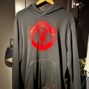 Manchester United svart hoodie - Svart hoodie med stor röd Manchester United-logga på. Perfekt för dig som vill visa vilket lag du håller på.