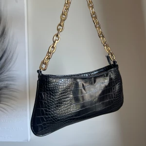 Svart axelväska med kedja - Snygg svart axelväska i croco-mönstrat syntetmaterial med en chunky guldfärgad kedja som axelrem. Väskan har en stilren, avlång form och glänsande finish som ger en lyxig vibe. Perfekt för dig som vill ha en trendig accessoar med statement-detaljer.