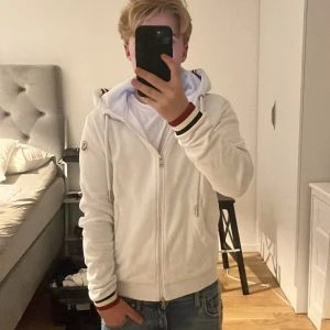 Vit hoodie från Moncler med dragkedja | S - Säljer en vit hoodie från Moncler med hel dragkedja och huva. Tröjan har Moncler-logga på ärmen och ribbade muddar med röd, vit och marinblå rand. Snygga detaljer med snörning i huvan och fickor med dragkedja. Perfekt för dig som gillar stilren och sportig look. Passar 165-175