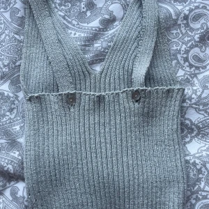 Grå ribbad stickad linnetopp - Säljer en grå ribbad stickad linnetopp med breda axelband och v-ringning. Toppen har dekorativa knappar framtill och ett diskret hålmönster på baksidan. Perfekt för att skapa en chill och trendig look. Den är elastisk så den ser mindre ut en vad den är.