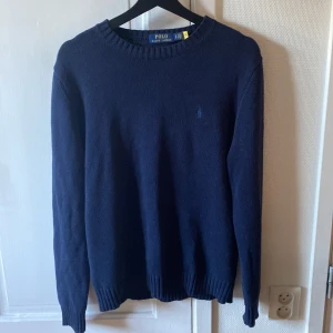 Polo Ralph Lauren mörkblå stickad tröja( NYSKICK) - Klassisk mörkblå stickad tröja från Polo Ralph Lauren med rund halsringning och diskret broderad logga på bröstet. Tröjan har ribbade muddar vid ärmslut och nederkant, perfekt för en stilren och enkel look. Priset är ej hugget i sten, skick är väldigt bra använd fåtal gånger. Vid Interesse eller något annat är de bara att höra av er
