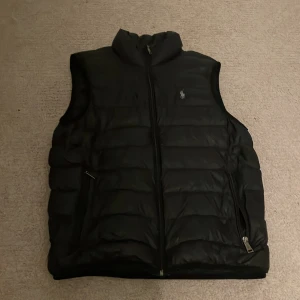 Svart dunväst från Polo Ralph Lauren - Svart dunväst från Polo Ralph Lauren med klassisk ståkrage och broderad logga på bröstet. Västen har två sidofickor med dragkedja och en kraftig dragkedja framtill. Den quiltade designen ger en snygg och sportig look, perfekt för lager-på-lager.