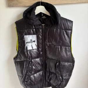 Svart dunväst med huva från H&M Divided - Cool svart dunväst från H&M Divided med glansig yta och tryck på bröstet med texten 'Voice Poison'. Västen har huva i sweatshirtmaterial, två fickor med knappstängning och dragkedja framtill. Insidan har en neonfärgad detalj längs kanten.