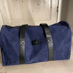 Lanvin Paris weekendbag navy blå mocka - Säljer en snygg och rymlig weekendbag från Lanvin i mörkblå mocka med svarta skinnhandtag. Väskan har dragkedja upptill och stilren design med Lanvin-logga framtill. Perfekt för resor eller gymmet när du vill ha lite extra stil.