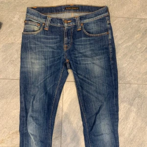 Blå jeans från Nudie Jeans - Snygga blå jeans från Nudie Jeans med klassisk femficksdesign och orange sömmar. Jeansen har en normal passform och är tillverkade i slitstarkt denim. Perfekta för en avslappnad stil. De är i storlek W26 men sitter mer som W27! Väldigt bra skick inga defekter alls!