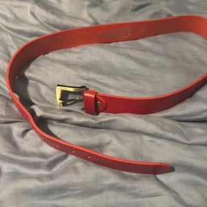 Rött skärp i skinn från KMG - Snyggt rött bälte från KMG i äkta skinn med klassiskt spänne i metall. Bältet har en stilren design utan mönster och passar perfekt till jeans eller kostymbyxor. Märket är präglat på insidan av bältet.