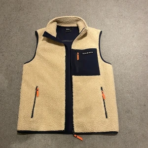 Beige fleeceväst från Ocean Works - Säljer en beige fleeceväst från Ocean Works i storlek S. Västen har marinblå detaljer, tre fickor med orange dragkedjor och hög krage. Perfekt att slänga över en hoodie för extra värme. Enkel och clean design med en bröstficka framtill. Nästan inte använd!!