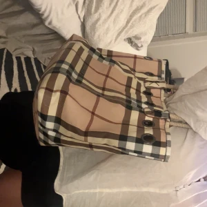 Rutig shoppingväska från Burberry - Säljer en klassisk shoppingväska från Burberry med det ikoniska rutiga mönstret i beige, brunt, svart och vitt. Väskan har grova rep-handtag och metallögla-detaljer upptill. Perfekt storlek för att få plats med allt du behöver och riktigt snygg att bära på axeln.