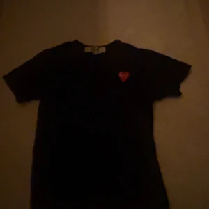 Svart Comme des Garçons Play t-shirt - Svart t-shirt från Comme des Garçons Play i storlek S. Klassisk modell med korta ärmar och rund hals. På bröstet sitter det ikoniska röda hjärtat med ögon, broderat som en patch. T-shirten är gjord i mjuk bomull och har en clean, stilren vibe.