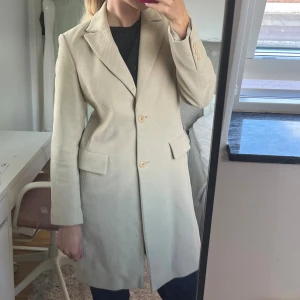Beige trenchcoat  - Stilren beige kappa från Turnover med klassisk krage, knappar framtill och fickor.Jackan är tillverkad i manchestertyg och har en rak, elegant passform som funkar till många looks. Perfekt för dig som gillar en clean och tidlös vibe.