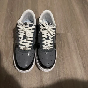 BAPE STA sneakers svart/grå - Snygga BAPE STA sneakers i svart och grått med vit sula och vita skosnören. Skorna har en glansig finish i syntetmaterial och den ikoniska stjärnan på sidan. Perfekt för dig som vill sticka ut med streetwear-vibe.