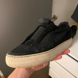 Svarta sneakers i mocka med vit sula - Stilrena svarta sneakers i mocka med tjock vit platt sula. Skon har en minimalistisk design utan synlig snörning och en rund tå. Perfekta för dig som gillar en clean och modern look.Dem har en skada i hälen men den påverkar inte något och ingeting man tänker på.