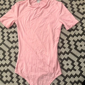 Ljusrosa bodysuit med kort ärm - En enkel och stilren ljusrosa bodysuit med korta ärmar och rund hals. Gjord i mjuk bomull som känns skön mot huden. Perfekt att styla med jeans eller kjol för en clean look. Bodyn har klassisk passform och är lätt att matcha. Aldrig använd, endast legat i garderoben.