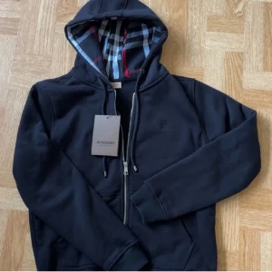 Svart hoodie från Burberry - Snygg svart hoodie från Burberry med dragkedja och klassiskt rutigt mönster i huvan. Tröjan har långa ärmar och ribbade muddar. Perfekt för en stilren look.