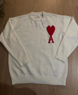 Vit stickad tröja med rött hjärta från Ami - Vit stickad tröja från Ami Alexandre Mattiussi med rund hals och ribbade muddar. På bröstet finns ett stort rött hjärta och bokstaven A. Tröjan har lång ärm och är tillverkad i mjuk ull, perfekt för dig som gillar clean design med en twist.
