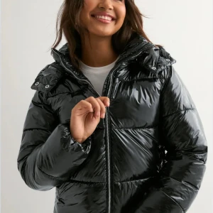 Svart pufferjacka från Nelly - Säljer en svart, glansig pufferjacka med huva och dragkedja framtill. Jackan är perfekt för kalla dagar. Materialet är syntet och jackan har långa ärmar. Använd endast en säsong och inte använt den så flitigt. Nypris 800kr, mitt pris kan diskuteras, kom privat för mer bilder🥰💞 (en av knapparna har lossnat på vänster fickan) 