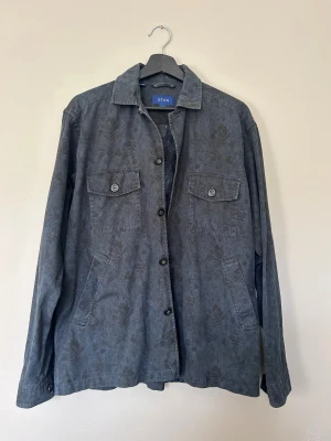 Mörkgrå overshirt från Eton, M - Snygg mörkgrå overshirt från Eton med diskret blommigt mönster. Jackan har två bröstfickor med knappar, sidfickor och klassisk krage. Tillverkad i bomull och stängs med knappar framtill. Perfekt lager-på-lager-plagg för en chill och stilren look.