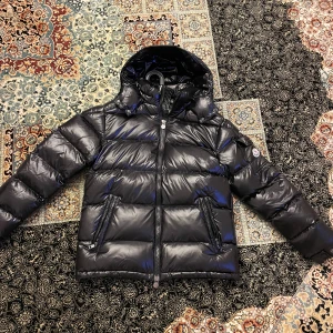 Svart Moncler pufferjacka med huva - Säljer en svart pufferjacka från Moncler med glansig finish, huva och två dragkedjefickor. Jackan har klassisk Moncler-logga på ärmen och quiltad design. Perfekt för kalla dagar och riktigt snygg streetstil.