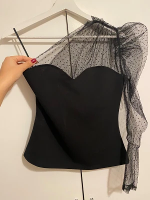 Svart one shoulder topp med meshärm - Säljer en svart topp med en axelbandslös design på ena sidan och en transparent meshärm med svarta prickar på andra sidan. Toppen har en figurnära passform och cool asymmetrisk look. Perfekt för dig som vill sticka ut med en unik och trendig stil.