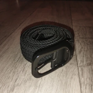 Svart flätad bälte med metallspänne - Snyggt svart bälte med flätad design och ett stilrent svart metallspänne. Bältet är flexibelt och har en modern look som passar till jeans eller chinos. Materialet är syntet och spännet ger en cool touch till outfiten.