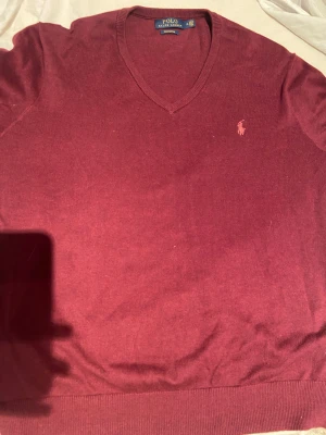 Vinröd v-ringad tröja Polo Ralph Lauren - Vinröd v-ringad tröja från Polo Ralph Lauren med klassisk broderad logga på bröstet. Tröjan är stickad i mjuk bomull och har ribbade muddar vid ärmslut och nederkant. Perfekt för dig som gillar stilrena och tidlösa plagg.