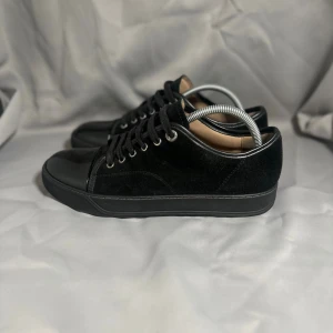 Lanvins  - Snygga svarta sneakers med ovandel i mjuk mocka och detaljer i blankt lack på tå och häl. Skorna har rund tå, platt sula och snörning med metallöglor. Perfekt för dig som gillar stilrena och moderna skor med lite extra edge.