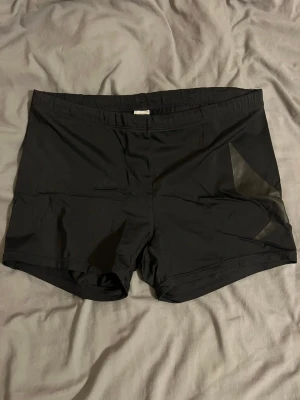 Svarta träningsshorts från SOC - Svarta träningsshorts från SOC i storlek M. De har en enkel design med diskreta svarta detaljer på sidorna och är tillverkade i ett syntetmaterial som torkar snabbt. Passformen är normal och midjan är midwaist. Perfekta för gymmet eller löprundan.