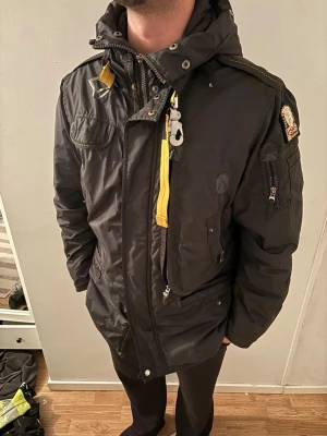 Svart Parajumpers jacka med huva - Svart Parajumpers jacka med ikonisk gul rem och patch på ärmen. Jackan har flera fickor, dragkedjor och en hög krage med huva. Tillverkad i slitstark polyester och har coola detaljer som metallspänne och robust känsla.