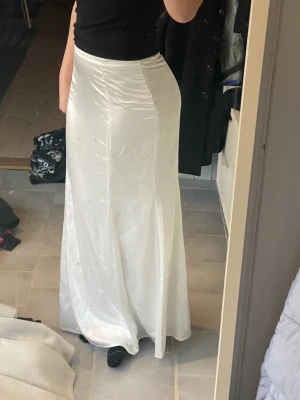 Vit långkjol i satin från H&M - Säljer en vit, glansig långkjol från H&M i mjukt satinmaterial. Kjolen har en figurnära passform upptill och faller snyggt ner mot golvet. Aldrig använd och prislappen kvar 