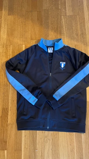 Malmö FF blå träningströja - Mörkblå officiell träningsmerch från Malmö FF med ljusblå detaljer på krage och ärmar. Jackan har dragkedja framtill och klubbens broderade logga på bröstet. Tillverkad i polyester, perfekt för fotboll och sportiga dagar. Nypris: 899 Mitt pris: 299