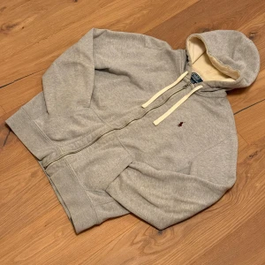 Grå hoodie från Polo Ralph Lauren - Grå hoodie från Polo Ralph Lauren med dragkedja och vit snörning i huvan. Tröjan har ett broderat rött Polo-logo på bröstet, två fickor fram och mjukt, beige foder i huvan. Perfekt för chill dagar och enkel att matcha med jeans eller joggers.