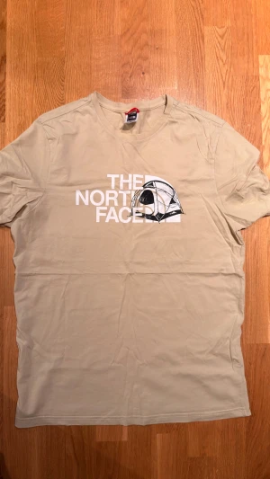 Beige t-shirt från The North Face - Snygg beige t-shirt från The North Face med vit logga och ett coolt tältmotiv på bröstet. Klassisk passform med rund hals och korta ärmar. Tillverkad i mjuk bomull som är skön att ha på sig hela dagen.