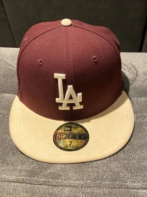 Vinröd LA Dodgers keps New Era 59FIFTY - Snygg LA Dodgers 59FIFTY keps från New Era i vinrött med beige skärm och detaljer. Framtill broderat LA-logo i vitt, på sidan broderad Dodger Stadium emblem. Klassisk flatbrim och MLB-logga bak. Perfekt för dig som gillar streetstyle och baseball.
