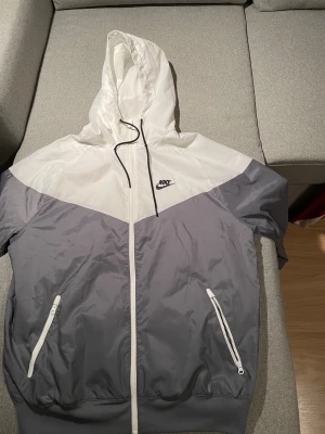 Nike vindjacka i vitt och grått - Nike vindjacka med huva i vitt och grått. Jackan har dragkedja framtill, två fickor med dragkedja och svart snörning vid huvan. Tillverkad i lätt polyester med meshfoder för extra komfort. Perfekt för dig som gillar sportig och clean stil.