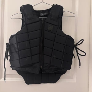 Svart ridväst Kids L med snörning - Svart quiltad ridväst från Tekna i storlek Kids L. Västen har dragkedja framtill, snörning i sidorna och vadderad design för extra skydd. Perfekt för dig som vill ha en cool och funktionell look i stallet.