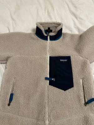Beige teddyjacka från Patagonia S - Mysig beige teddyjacka från Patagonia med marinblå detaljer och hög krage. Jackan har dragkedja framtill, två sidofickor med dragkedja och en bröstficka i marinblått tyg. Perfekt för kalla dagar och riktigt skön att bära.