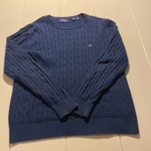 Marinblå kabelstickad tröja GANT - Snygg marinblå kabelstickad tröja från GANT med rund hals och diskret broderad logga på bröstet. Tröjan har långa ärmar och ribbade muddar vid ärmslut och nederkant. Perfekt för dig som gillar klassisk och stilren look.